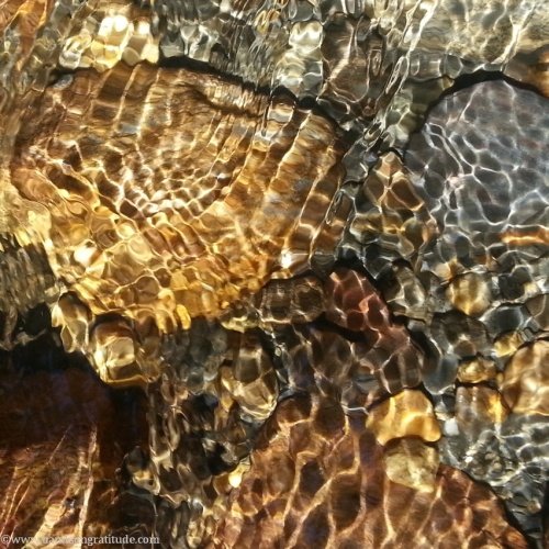 Macro photo of sun reflecting on moving water over golden and grey rocks. It is only my world I create, and you, yours. Be well. You are dear in the eyes of the Universe.